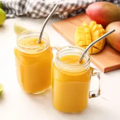 Dwa orzeźwiające drinki z mango w słoikach z metalowymi słomkami, obok świeżych mango i limonek.