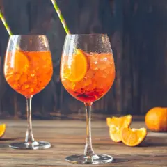 Dwa kieliszki Aperol Spritz z plasterkami pomarańczy i słomkami na drewnianym stole.