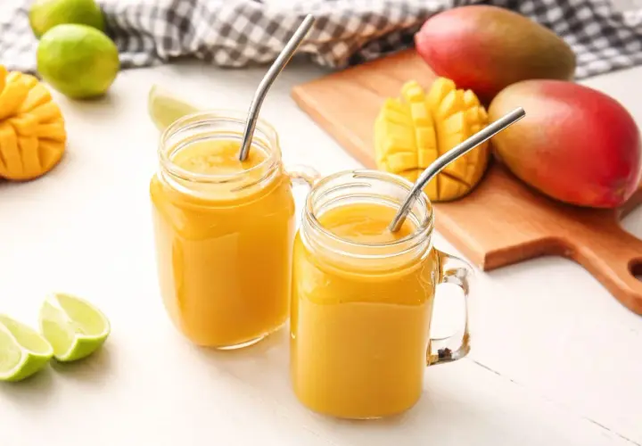 Dwa orzeźwiające drinki z mango w słoikach z metalowymi słomkami, obok świeżych mango i limonek.