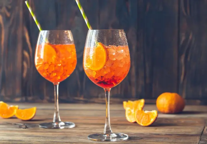 Dwa kieliszki Aperol Spritz z plasterkami pomarańczy i słomkami na drewnianym stole.